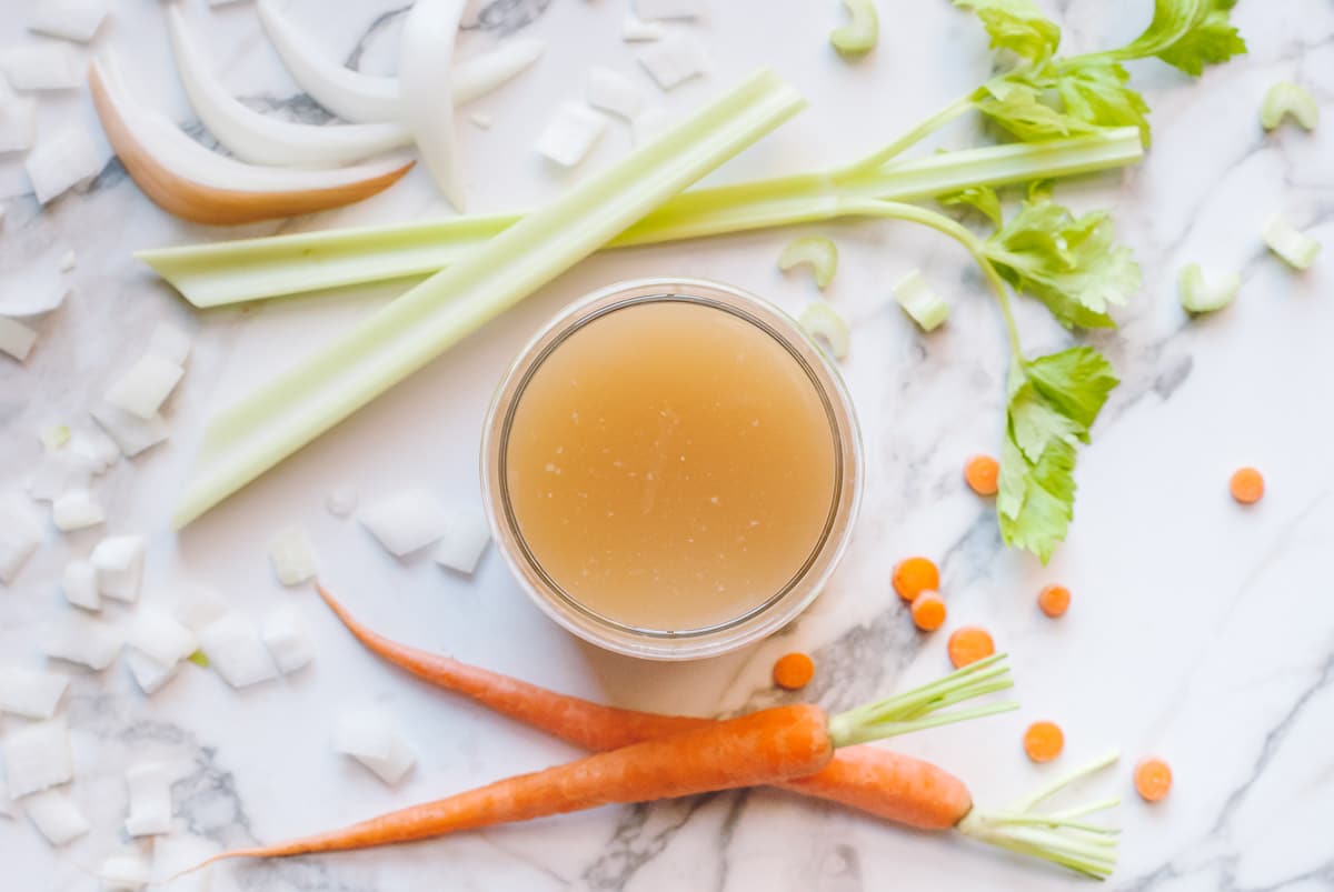 Over head shot of a cup of broth with celery sticks, carrots, and onions scattered around.